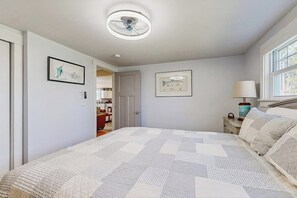 A bedroom with a bed, a ceiling fan, a window, and a door leading to a bathroom. - Coastal bungalow with large yard, beautiful kitchen, & great location (Boothbay Harbor)