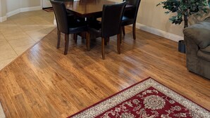 Un coin repas avec une table et des chaises en bois, un tapis à motifs rouges, une plante en pot et une fenêtre avec des stores.
