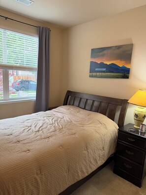 Un dormitorio con cama, mesita de noche con lámpara, cuadro en la pared y ventana con vistas a un coche y vegetación.