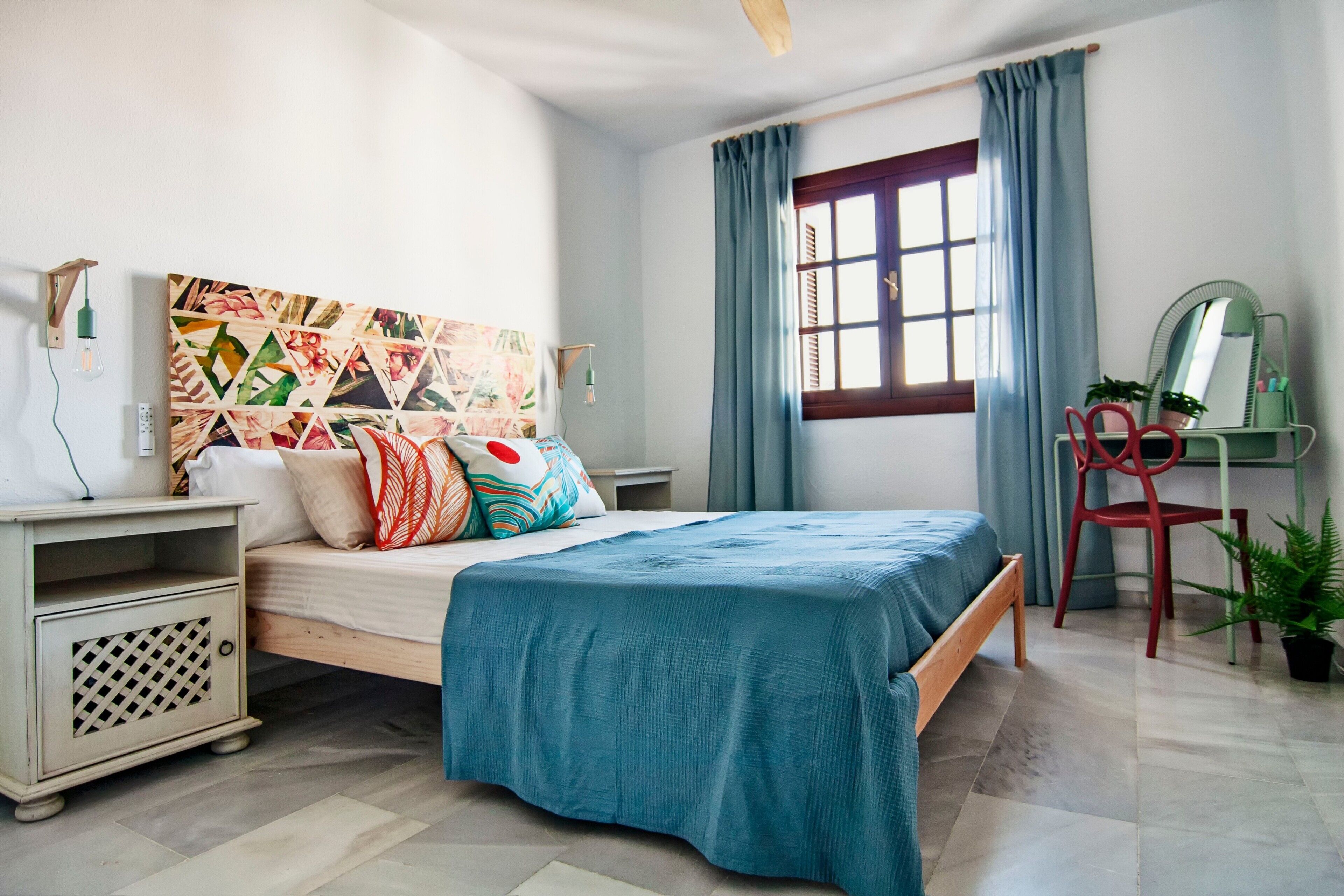 A bedroom with a bed, a nightstand, a desk with a chair, a window with curtains, and a wall with a geometric painting.
