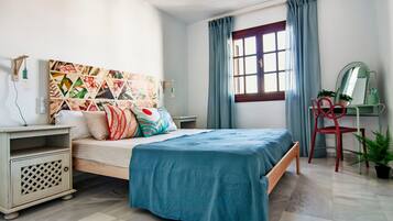 A bedroom with a bed, a nightstand, a desk with a chair, a window with curtains, and a wall with a geometric painting.