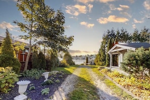 Ein Gartenweg führt zu einem Haus mit Blick auf ein Gewässer und eine Stadtlandschaft im Hintergrund.