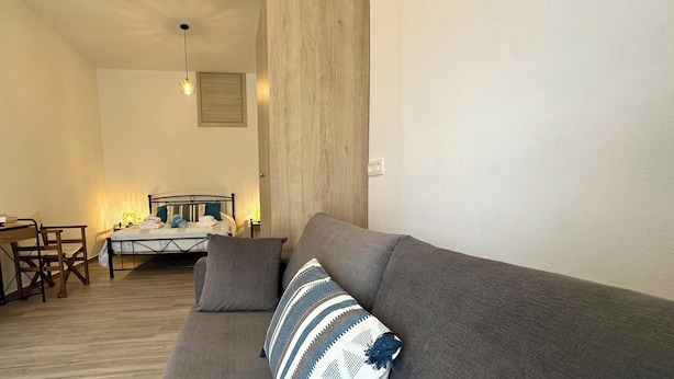 A bedroom with a bed, a grey sofa, a wooden floor, a desk, and a chair. - Black Sand Santorini Studio (Perissa)