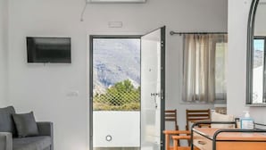 A modern room with a grey sofa, a TV mounted on the wall, a dining table with chairs, and a view of mountains through a glass door. - Black Sand Santorini Studio (Perissa)
