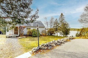 Ein kleines Haus mit Veranda, Steinmauer und einem See im Hintergrund.