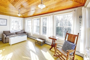 Ein Zimmer mit Holzboden, großen Fenstern, einer Couch, einem Couchtisch, einem Schaukelstuhl und einem kleinen Tisch.