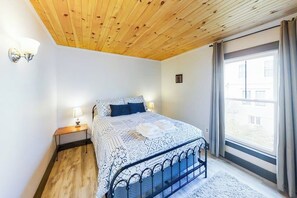 A bedroom with a wooden ceiling, a bed with patterned bedding, a nightstand with a lamp, and a window with curtains. - Newly Renovated Haven w/ Deck, Walk to Shops, Dining &  Marina, Pet friendly (Boothbay Harbor)