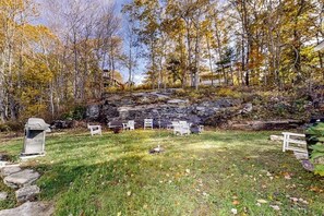 A natural outdoor setting with a grassy area, white outdoor furniture, and a rocky terrain. - Boothbay Cottage- Coastal home with new, full kitchen, walkable downtown location (Boothbay Harbor)