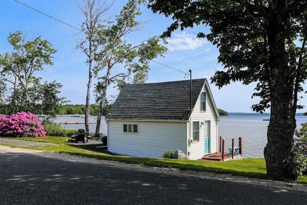 A small white cabin with a dark roof, a porch with a table and chairs, and a view of a lake.