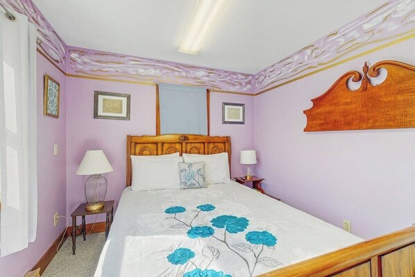 Quarto com cabeceira de madeira, cama com colcha floral, duas luminárias de cabeceira e quadros nas paredes.