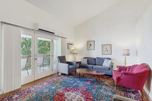 Ein Wohnzimmer mit einem blauen Sofa, einem rosa Sessel, einem gemusterten Teppich und einer Glastür, die zu einem Außenbereich führt.