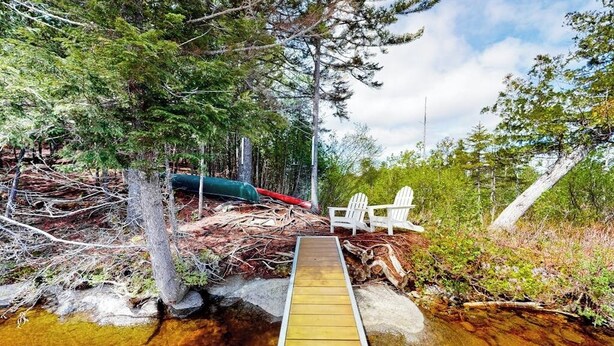 A wooden deck leading to a body of water, surrounded by trees and a white chair.