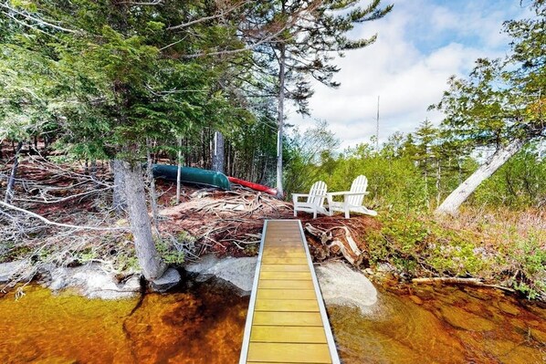 A wooden deck leading to a body of water, surrounded by trees and a white chair.