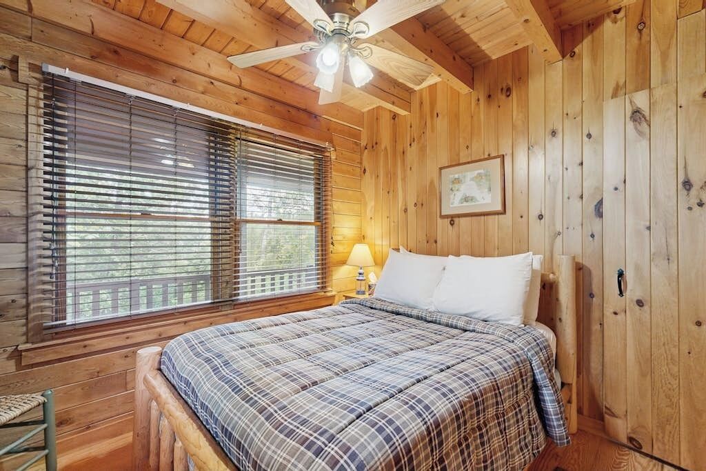 Un dormitorio de cabaña de madera con una cama, un ventilador de techo y una ventana con persianas.