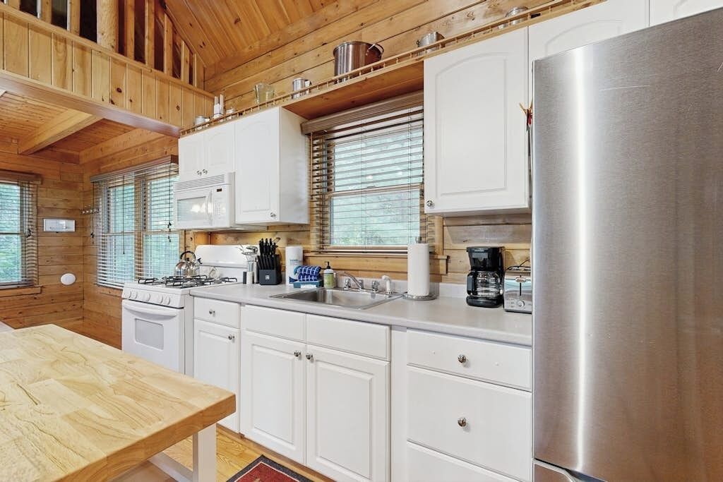 L’intérieur d’une cabane en bois comprend une cuisine équipée de meubles blancs, d’un réfrigérateur en acier inoxydable, d’une cuisinière et d’un évier.