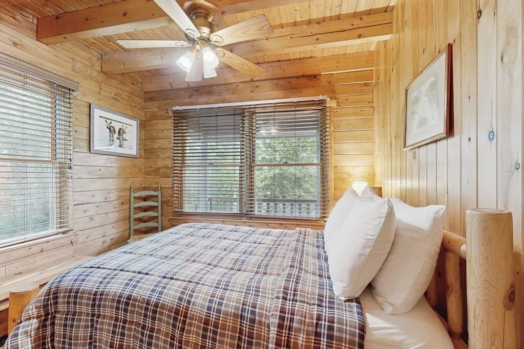 Un dormitorio de cabaña de madera con una cama, un ventilador de techo y dos ventanas con persianas.