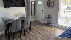 Uma área de refeições com televisão, cadeiras cinzas, uma pequena mesa com um vaso, uma porta com painel de vidro e uma janela com persianas.