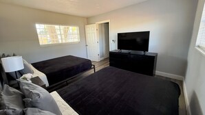 Quarto com duas camas, uma TV, uma cômoda, uma janela e uma porta.