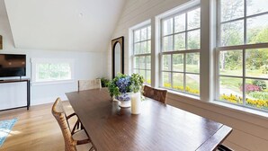 Een eethoek met een houten tafel, stoelen, een vaas met bloemen en grote ramen met uitzicht op groen.