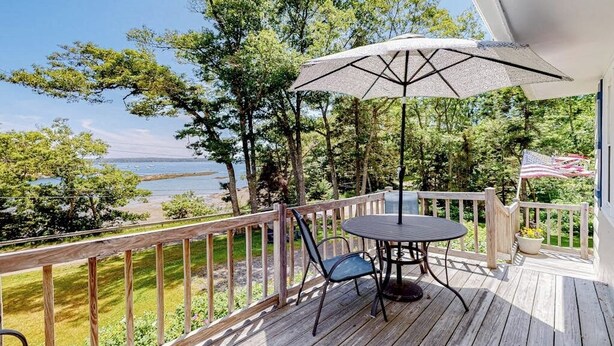 A wooden deck with a table and chairs, a large umbrella, and a view of the water. - Barrett's Watch Main House- Oceanfront on Lobster Cove with deck & shared firepit - walk to town (Boothbay Harbor)