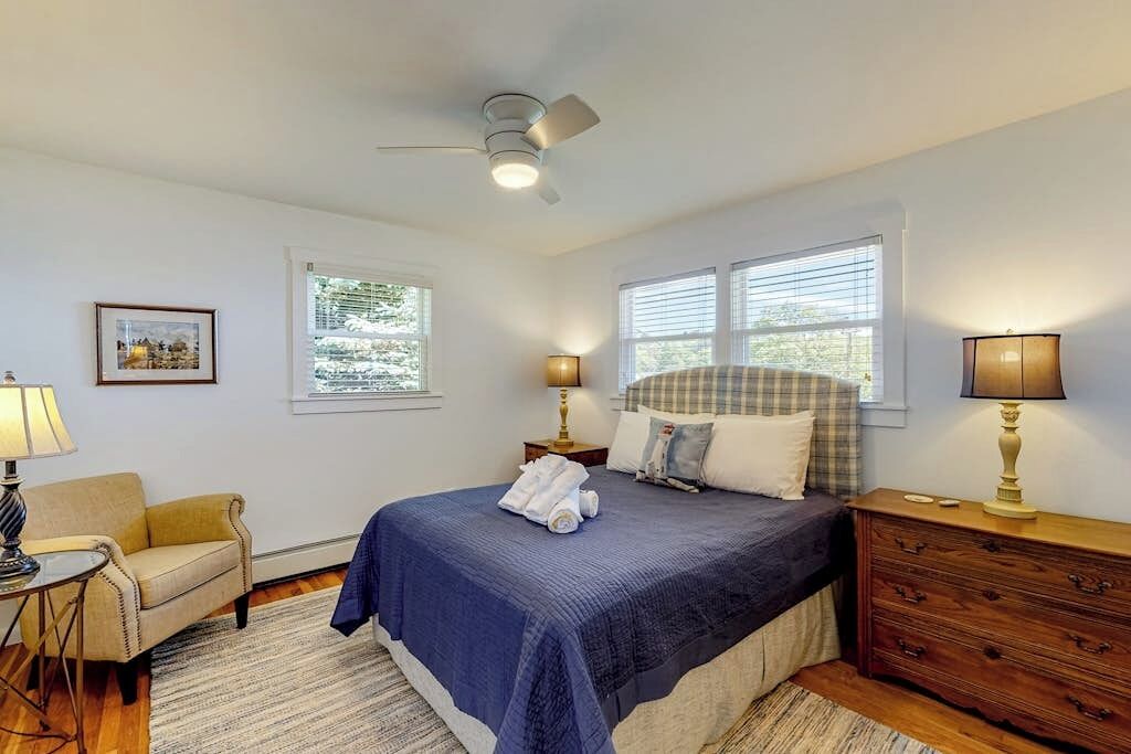 A bedroom with a bed, two bedside lamps, a ceiling fan, a window with blinds, a chair, a dresser, and a framed picture on the wall.