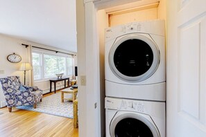 Ein Waschraum mit übereinander gestapelten Waschmaschinen und Trocknern, einem Holzboden, einem gemusterten Sofa und einem Fenster mit Jalousien.
