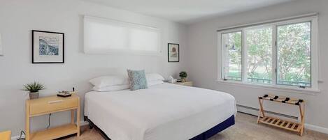 A bedroom with a bed, a bedside table with a plant, a window with a view, and a wall-mounted artwork.