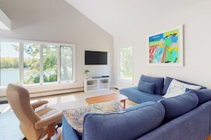 Un salón moderno con un ventanal grande, un sofá azul, un sillón beige, un televisor de pantalla plana y un cuadro colorido.