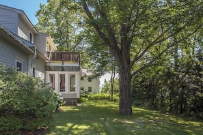 A two-story house with a visible balcony, surrounded by lush greenery and a large tree.