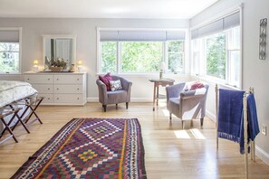 A bedroom with a bed, two chairs, a small table, a dresser, and a large window.