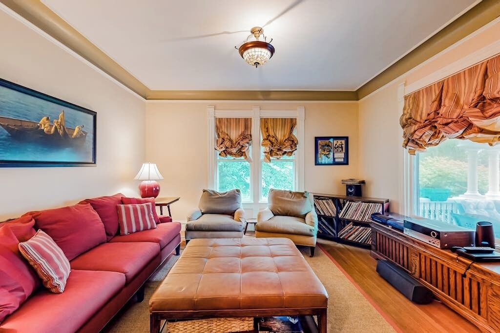 A living room with a red sofa, a brown ottoman, a wooden coffee table, a bookshelf, and a large window with curtains.