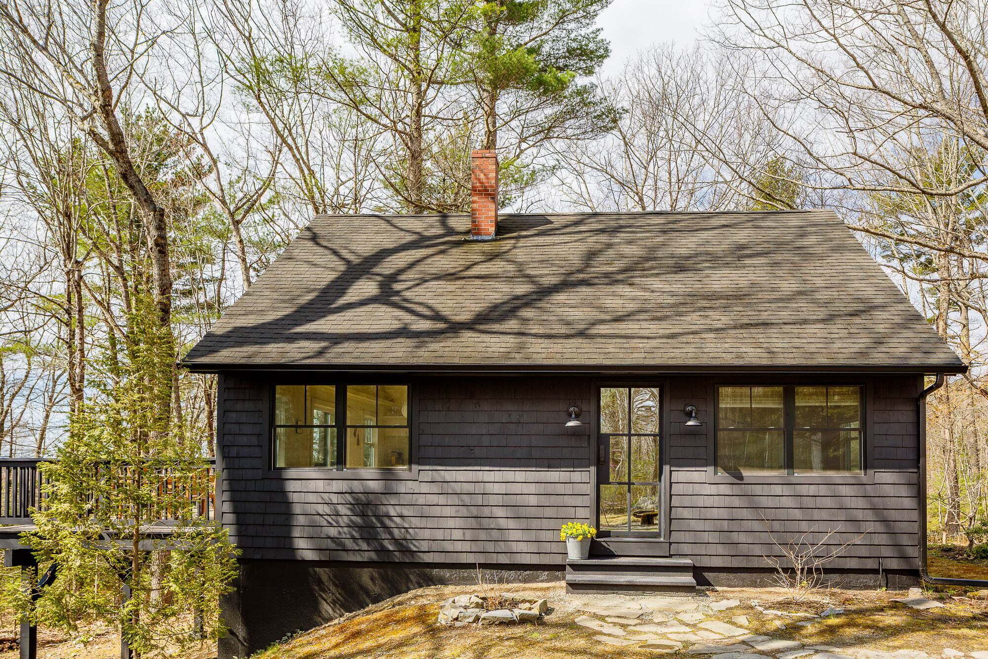 Une petite cabane aux couleurs sombres, dotée d’une cheminée, de grandes fenêtres et de fondations en pierre.