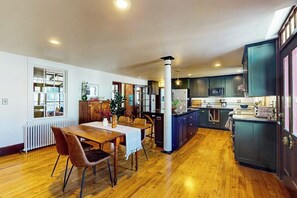 A spacious kitchen with dark cabinets, a central island, and a dining area with a wooden table and chairs. - Elegant Victorian Gothic Revival home with wet bar, sunroom, & washer/dryer (Belfast)
