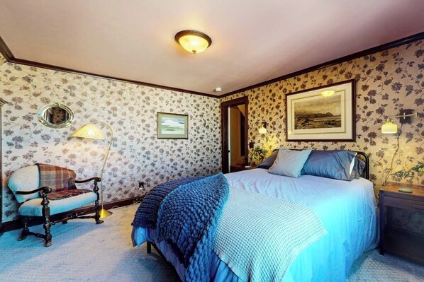 A bedroom with floral wallpaper, a bed with blue bedding, a vintage chair, and wall-mounted lamps. - Elegant Victorian Gothic Revival home with wet bar, sunroom, & washer/dryer (Belfast)