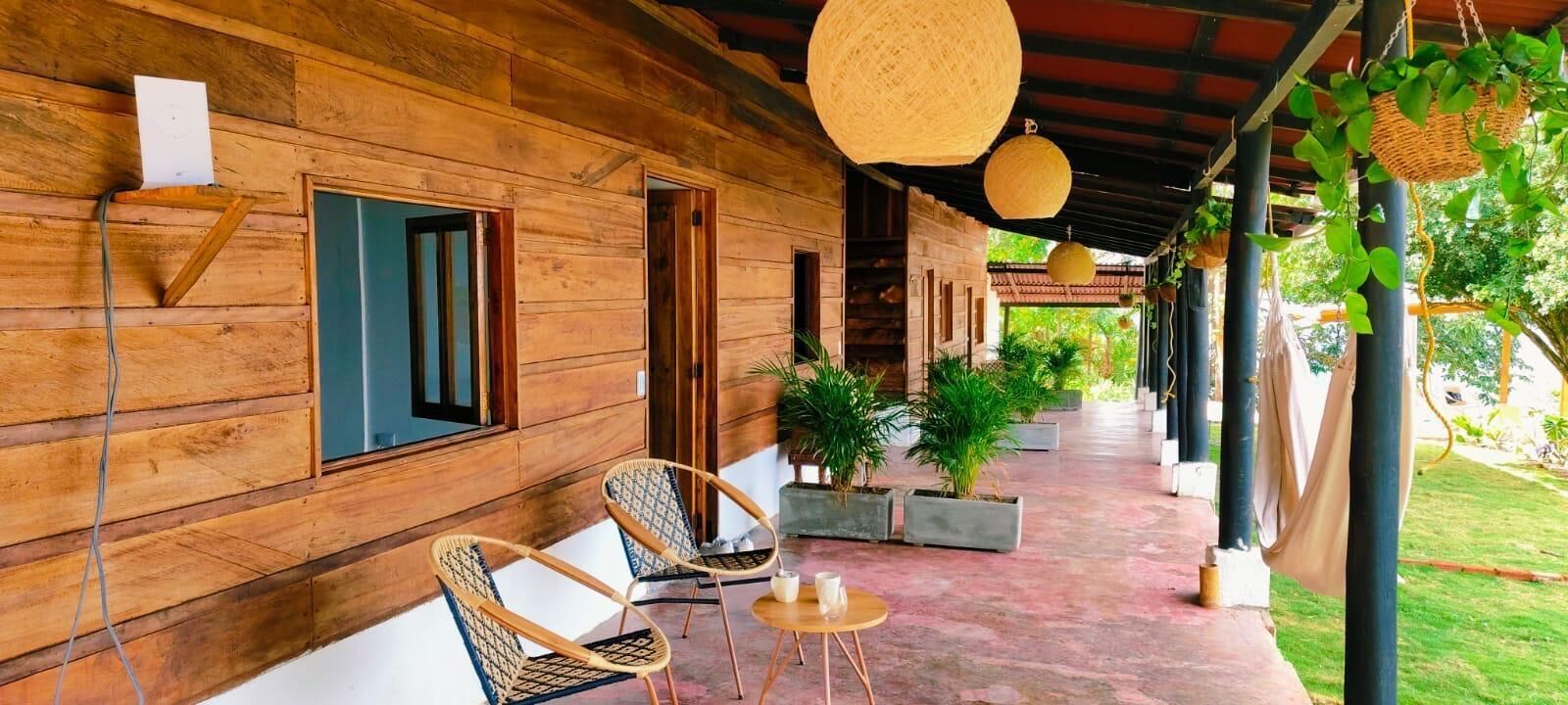 Uma cabana de madeira com varanda coberta, duas cadeiras de vime, uma mesa redonda pequena e plantas em vasos.