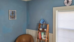 Ein Zimmer mit blauer Wand, einem Holzsessel, einem Bücherregal mit Büchern, einer dekorativen Anker-Figur und einer Wanduhr.