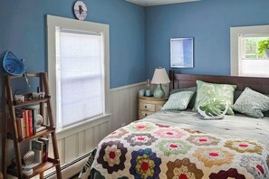 A bedroom with a bed, a nightstand, a lamp, a bookshelf, and a window with blinds.