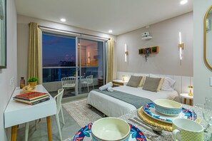 Een moderne hotelkamer met balkon, een bed met veel kussens, een nachtkastje met lamp, en een eethoek met een tafel gedekt voor twee personen.