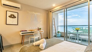 Ett modernt hotellrum med en säng, ett skrivbord, en stol, en balkong med utsikt över havet och en inramad tavla på väggen.
