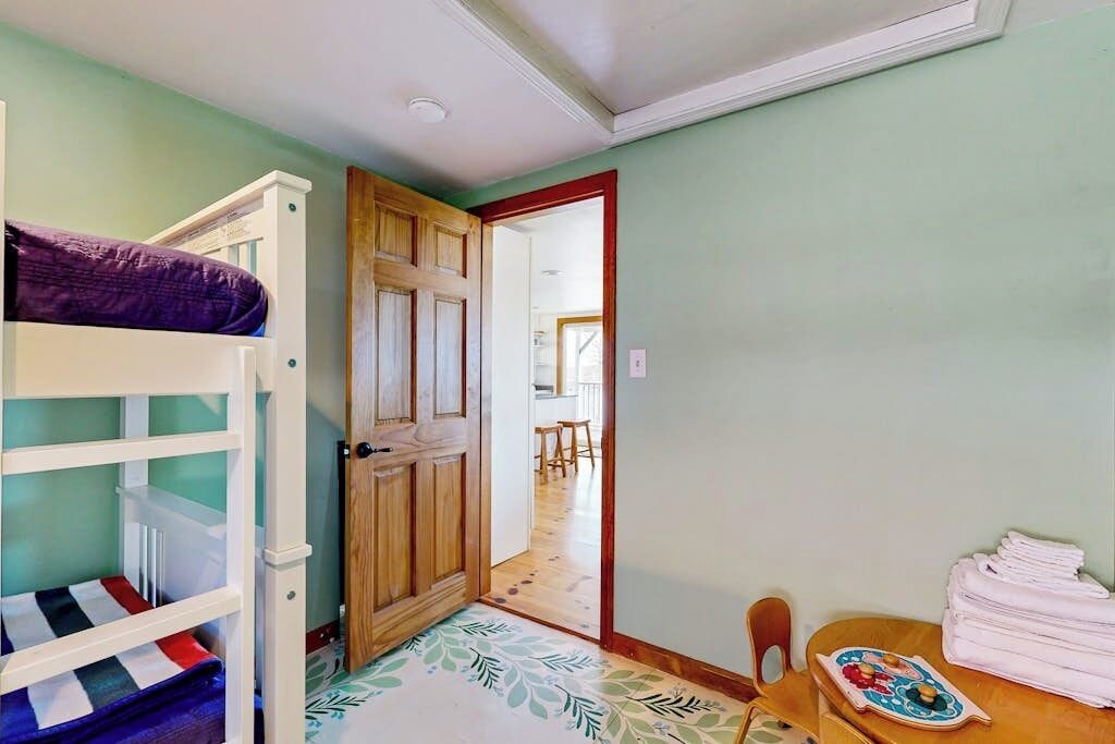 A room with bunk beds, a wooden door, a dining table with chairs, and a patterned rug.