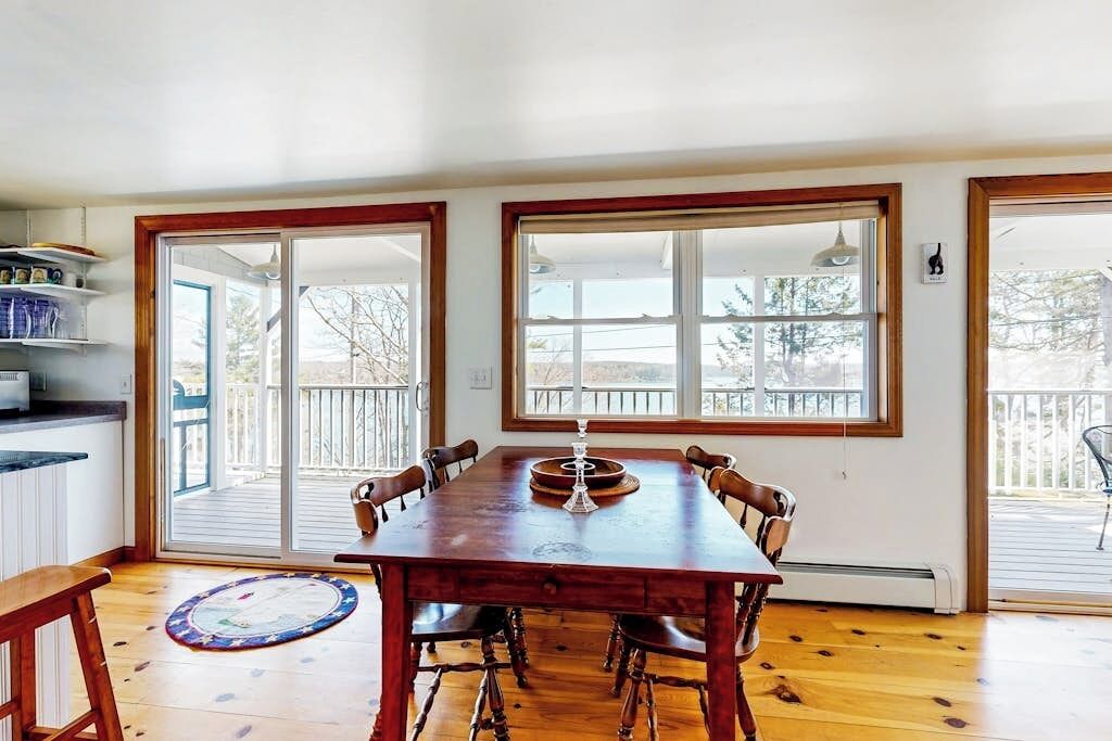 A dining area with a wooden table, chairs, and a view through large windows to an outdoor deck.