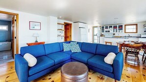 A living room with a blue sectional sofa, a round ottoman, and a dining area with wooden chairs and a table.