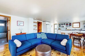 Ein Wohnzimmer mit einem blauen Ecksofa, einem runden Hocker und einem Essbereich mit Holzsesseln und einem Tisch.