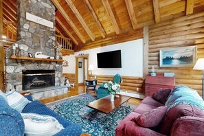 A cozy log cabin interior with a stone fireplace, wooden beams, a television, and a comfortable seating area.