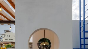 Een witte muur met een rond raam, waarachter een hanglamp te zien is en een gebouw met een geel bord.
