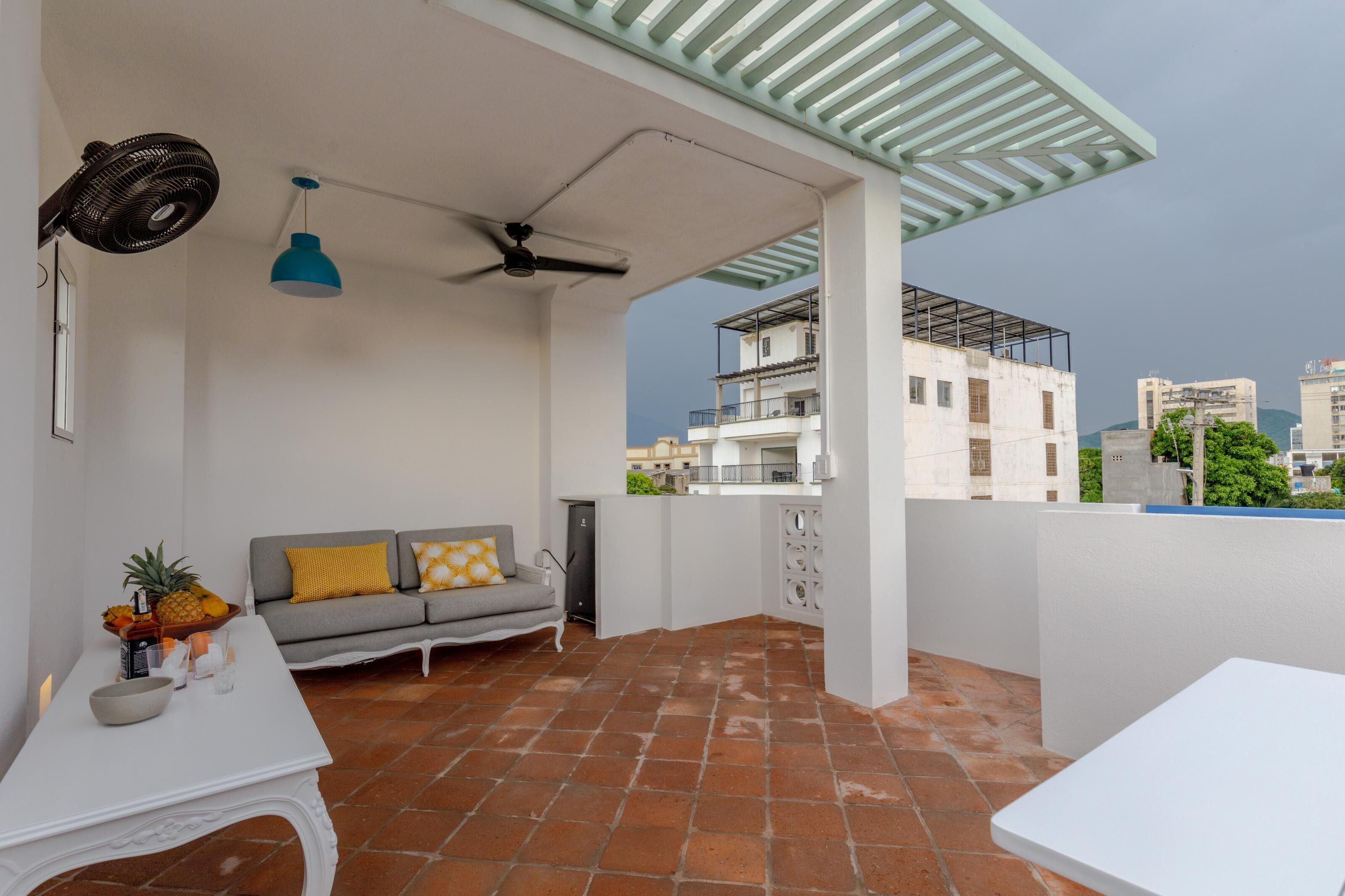 Ein Balkon mit weißer Couch, einem Tisch mit Obstschale, einer blauen Pendelleuchte und einem Deckenventilator.