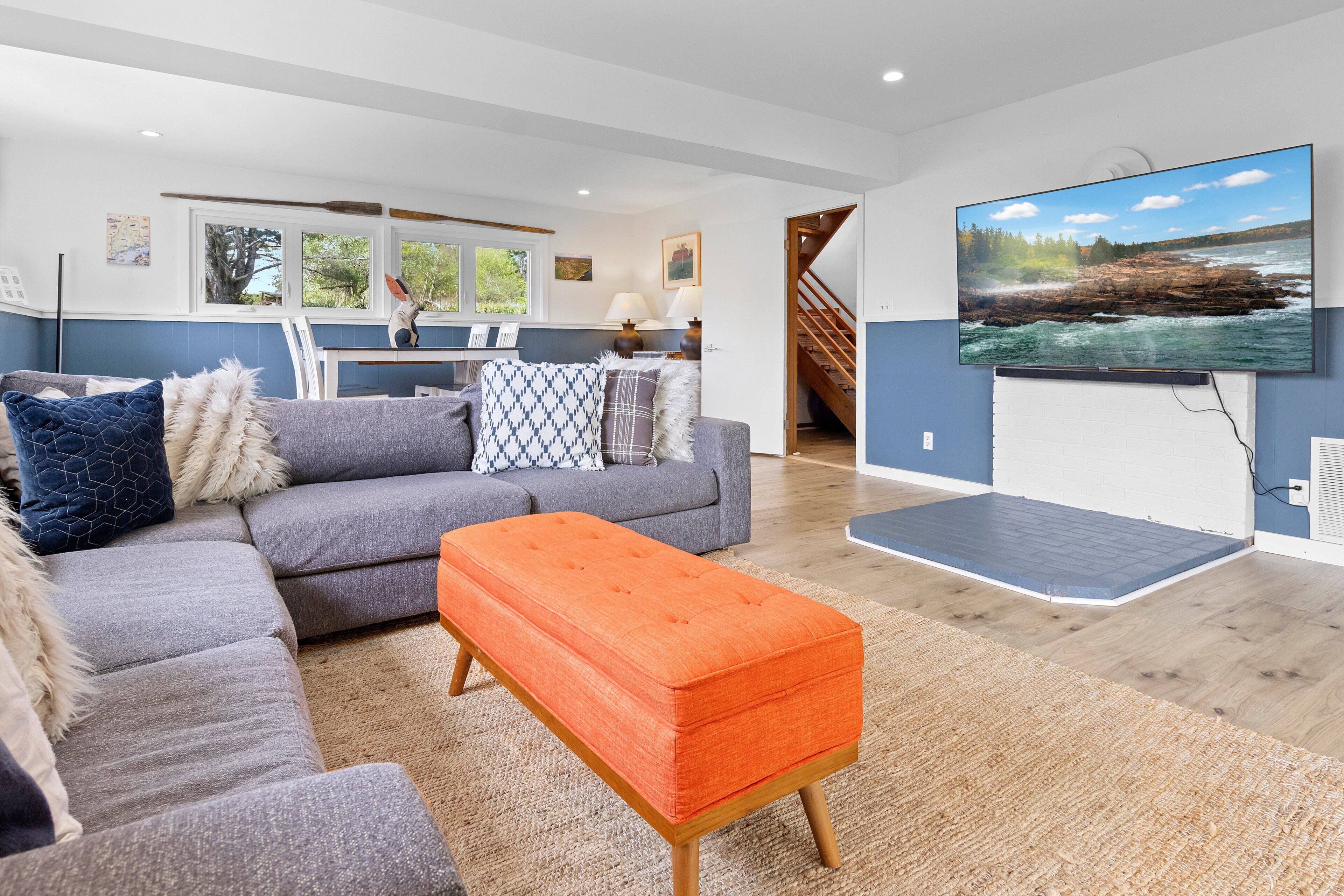 A modern living room with a sectional sofa, a bright orange ottoman, and a large flat-screen TV mounted on the wall.