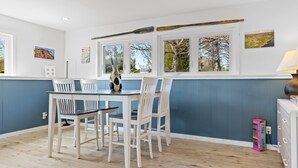 A dining area with a table and chairs, a window with a view of trees, and a lamp on a cabinet. - Evelyn's River Front Retreat- Spacious House with Modern Comforts and Nature (Cushing)
