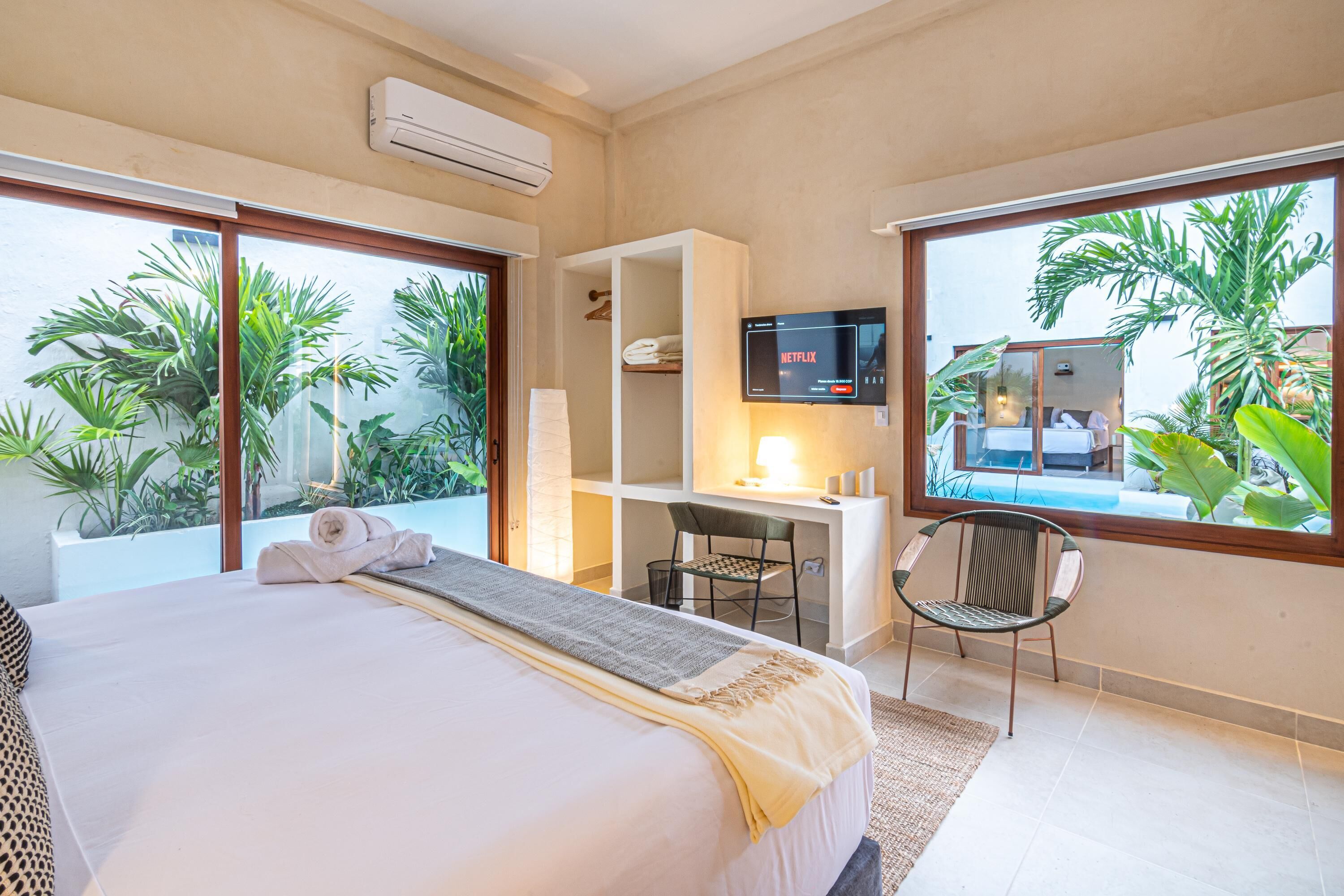 A hotel room with a bed, a desk, a TV, and large windows overlooking a garden.