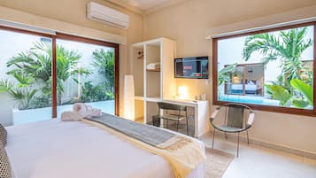 A hotel room with a bed, a desk, a TV, and large windows overlooking a garden.